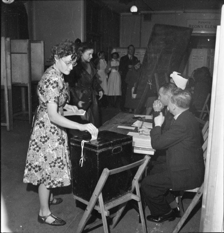 A woman places her ballot paper in a wooden ballot box watched by polling clerks at a table, 1945