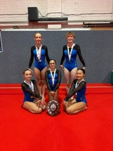 Five Dover Gymnastics Club gymnasts in blue and black leotards with medals, posing with the South East Bronze Level Team shield